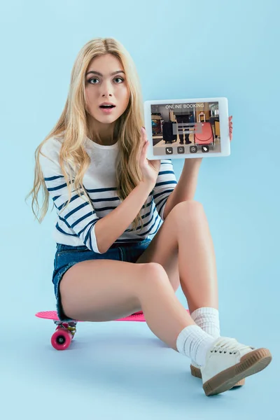 Menina loira incrível sentado no longboard e segurando tablet digital com aplicativo de reserva on-line na tela no fundo azul — Fotografia de Stock