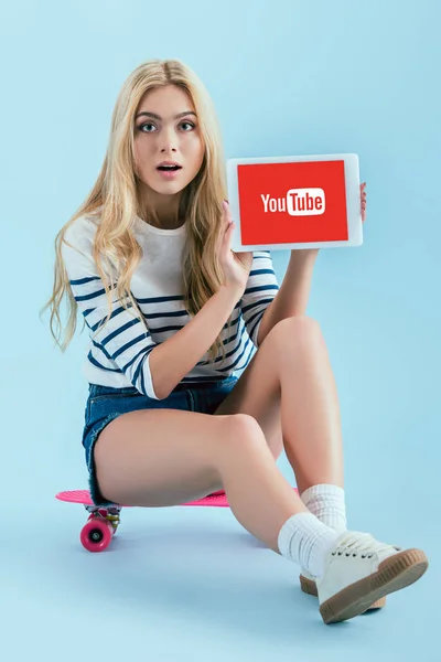 Amazed blonde woman sitting on longboard and holding digital tablet with youtube app on screen on blue background — Stock Photo