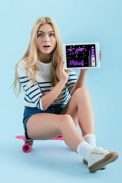 Erstauntes Mädchen zeigt digitales Tablet mit Business-App auf dem Bildschirm, während es auf Longboard auf blauem Hintergrund sitzt — Stockfoto