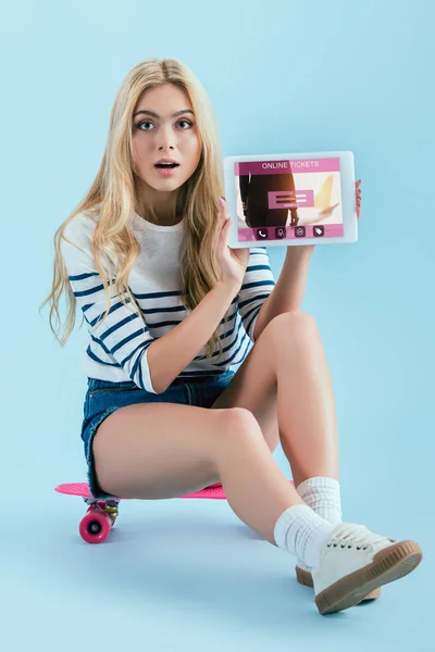 Sorpresa ragazza dai capelli lunghi che mostra tablet digitale con i biglietti online app sullo schermo e seduto sul longboard su sfondo blu — Foto stock