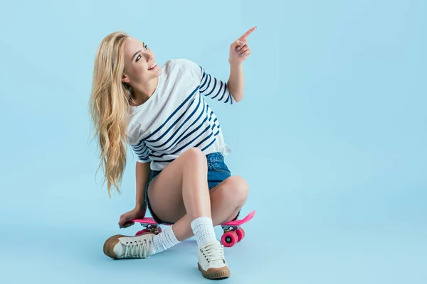 Joyeuse fille assise sur longboard rose et pointant du doigt sur fond bleu — Photo de stock