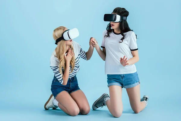 Chicas asustadas en VR auriculares de pie sobre las rodillas y cogidas de la mano sobre fondo azul - foto de stock