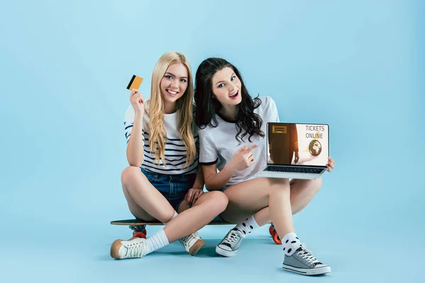 Mädchen auf Longboard halten Laptop mit Online-Tickets auf dem Bildschirm und Kreditkarte auf blauem Hintergrund — Stockfoto