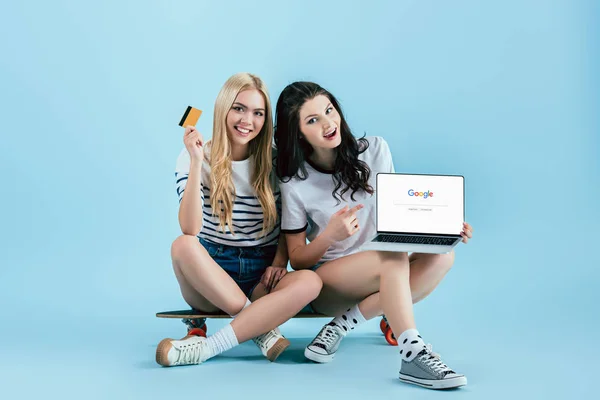 Stunning girls sitting on longboard and holding laptop with google website on screen and credit card on blue background — Stock Photo