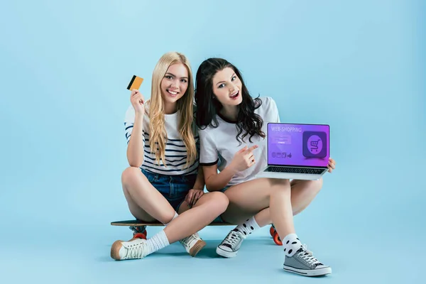 Meninas alegres no longboard segurando cartão de crédito e laptop com site de compras on-line na tela no fundo azul — Fotografia de Stock