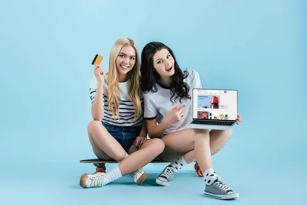 Studio shot of girls holding laptop with ebay website on screen and credit card while sitting on longboard on blue background — Stock Photo