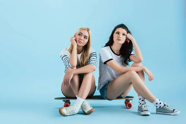 Chicas pensativas sentadas en longboard y caras apoyadas con las manos en el fondo azul - foto de stock