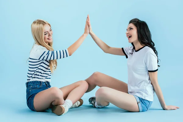 Chicas alegres en pantalones cortos de mezclilla posando en el suelo sobre fondo azul - foto de stock