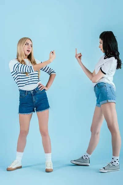 Studio shot of irritated girls showing middle fingers to each other on blue background — Stock Photo