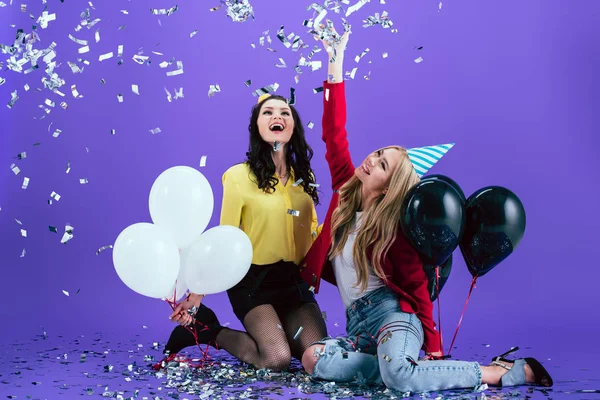 Riendo chicas en sombreros de fiesta sosteniendo globos y lanzando confeti sobre fondo púrpura - foto de stock