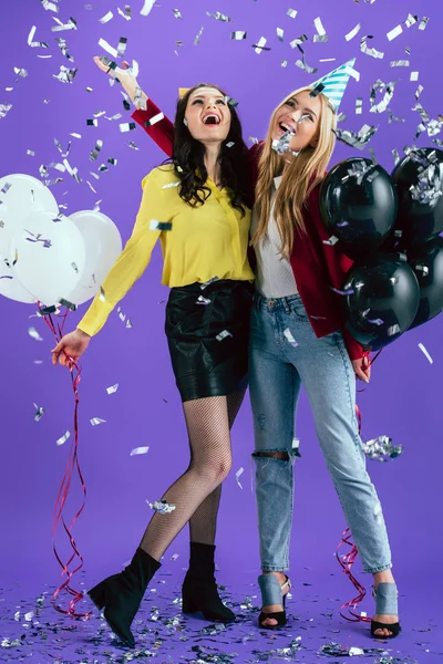 Chicas atractivas con globos riendo bajo confeti sobre fondo púrpura - foto de stock