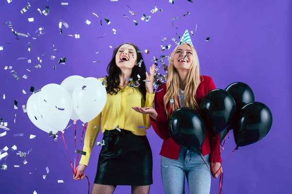Счастливые девушки с черно-белыми воздушными шарами позируют под конфетти на фиолетовом фоне — стоковое фото