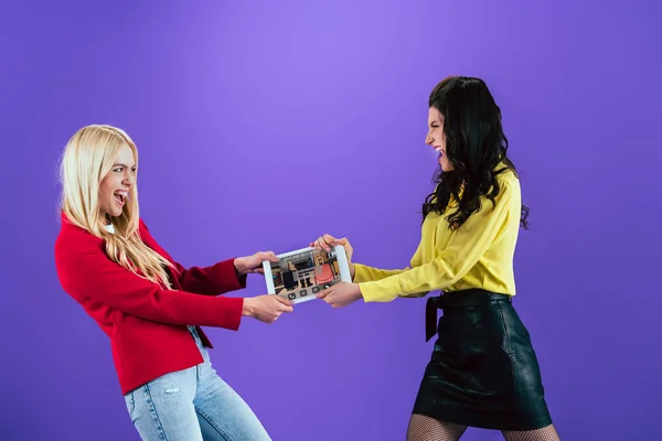 Chicas enojadas luchando por la tableta digital con aplicación de reserva en línea en la pantalla en el fondo púrpura - foto de stock