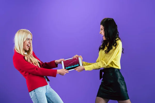 Jeunes femmes en colère se battant pour la tablette numérique avec l'application traiding à l'écran sur fond violet — Photo de stock
