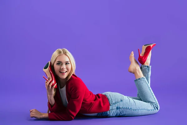Gorgeous smiling girl in red high-heeled shoes lying on floor and looking at camera on purple background — Stock Photo