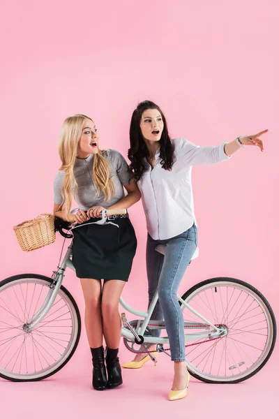 Surpris brunette fille pointant du doigt tout en posant sur le vélo avec un ami sur fond rose — Photo de stock