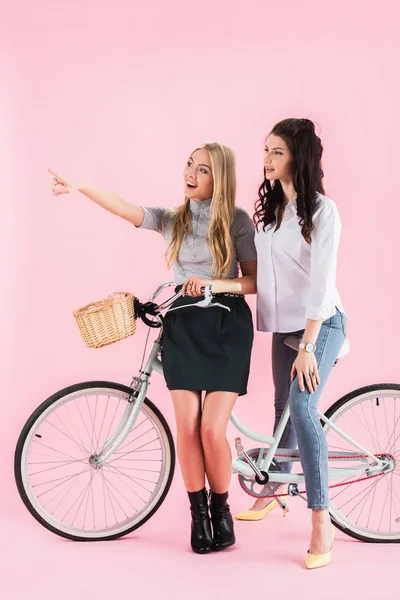 Rire fille blonde pointant du doigt tout en étant assis sur le vélo avec un ami sur fond rose — Photo de stock