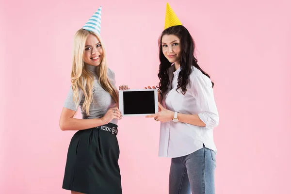 Чудові дівчата у вечірніх капелюхах тримають цифровий планшет з порожнім екраном ізольовані на рожевому — стокове фото