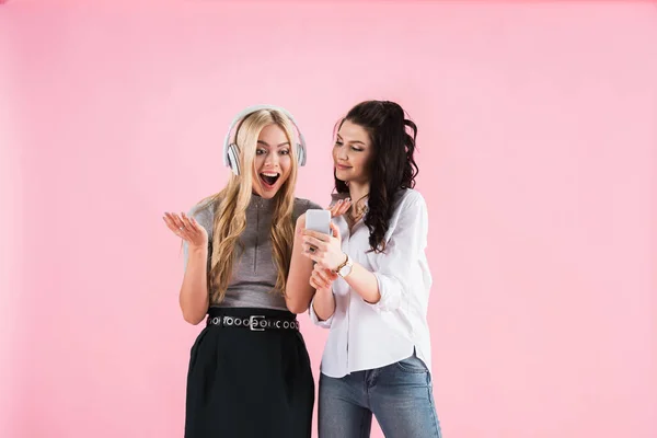 Meninas animadas bonitas com fones de ouvido e smartphones ouvindo música isolada no rosa — Fotografia de Stock