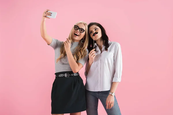 Chicas con bigote falso y gafas tomando selfie aislado en rosa - foto de stock