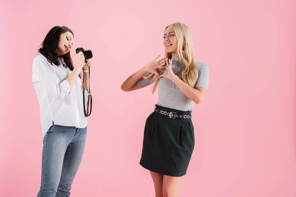 Chica rubia mostrando el signo del corazón mientras que amigo con la cámara tomando fotos aislado en rosa - foto de stock