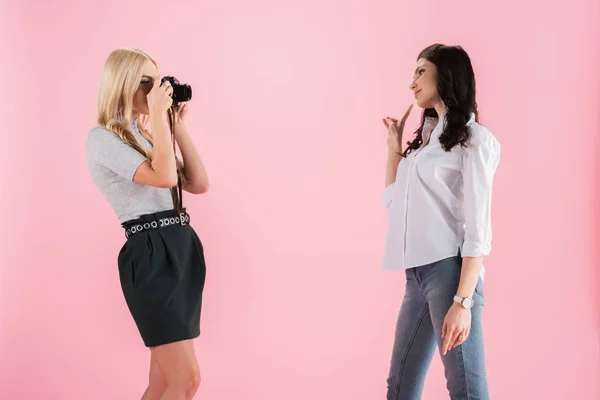 Блондинка фотографує, а дівчина брюнетка показує знак миру ізольовано на рожевому — стокове фото