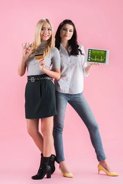 Chicas confiadas sosteniendo tarjeta de crédito y tableta digital con aplicación de fitness en la pantalla y mostrando el signo de aprobación en el fondo rosa - foto de stock