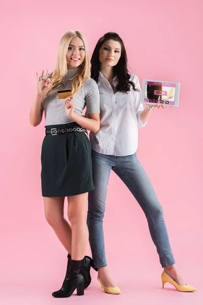 Chicas sonrientes posando con tarjeta de crédito y tableta digital con la aplicación de boletos en línea en la pantalla y mostrando el signo de aprobación en el fondo rosa - foto de stock