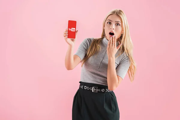 Chica impactada atractiva mostrando teléfono inteligente con aplicación de youtube en la pantalla, aislado en rosa - foto de stock