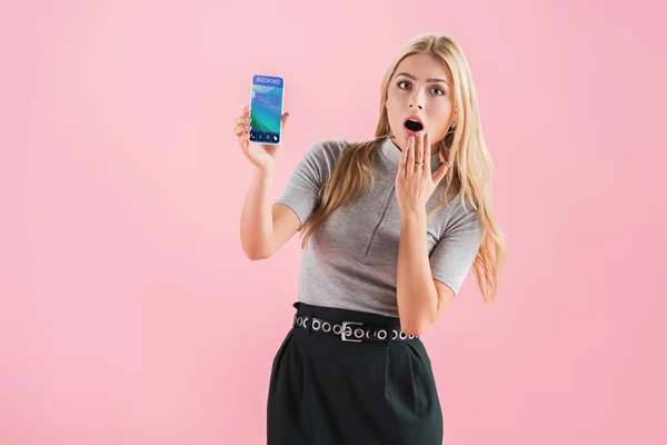 Schockierte Frau zeigt Smartphone mit Buchungs-App auf dem Bildschirm, isoliert auf rosa — Stockfoto