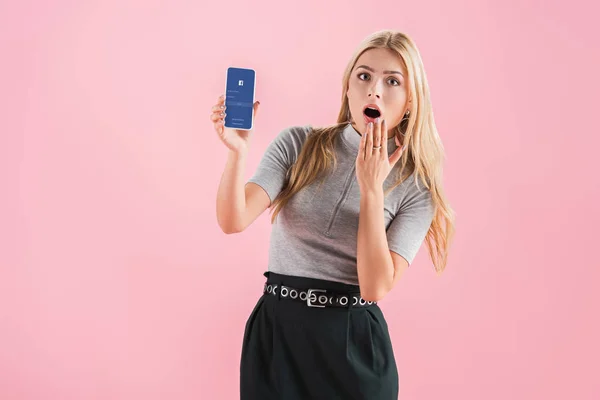 Chica impactada atractiva mostrando teléfono inteligente con aplicación de Facebook en la pantalla, aislado en rosa - foto de stock