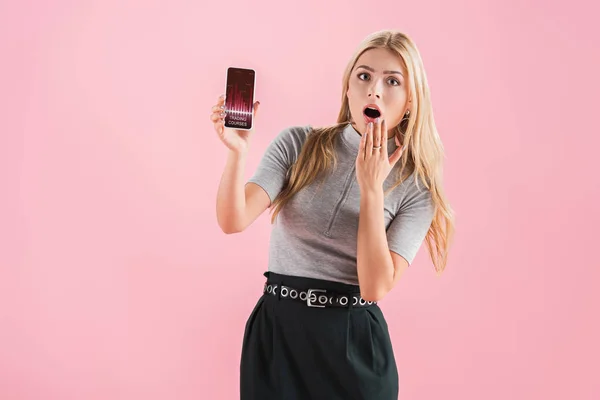 Mujer rubia impactada mostrando teléfono inteligente con cursos de comercio y gráfico en la pantalla, aislado en rosa - foto de stock