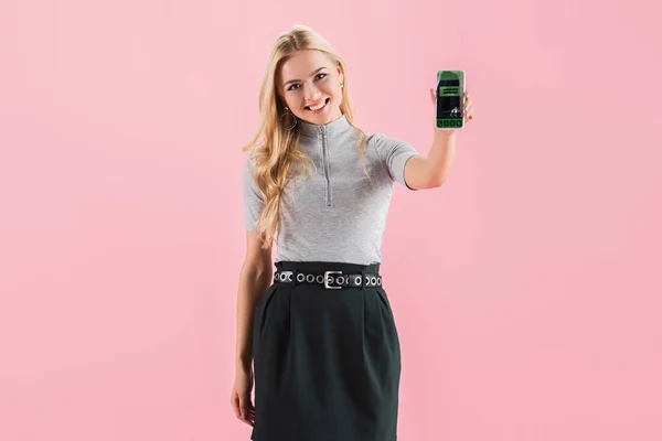 Smiling blonde girl showing smartphone with booking app on screen, isolated on pink — Stock Photo