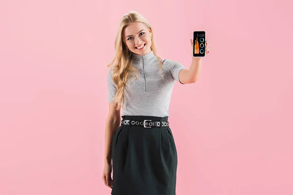 Lächelndes blondes Mädchen zeigt Smartphone mit Grafiken auf dem Bildschirm, isoliert auf rosa — Stockfoto