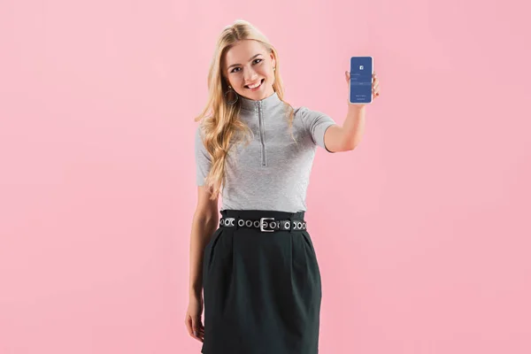 Cheerful blonde girl showing smartphone with facebook app on screen, isolated on pink — Stock Photo