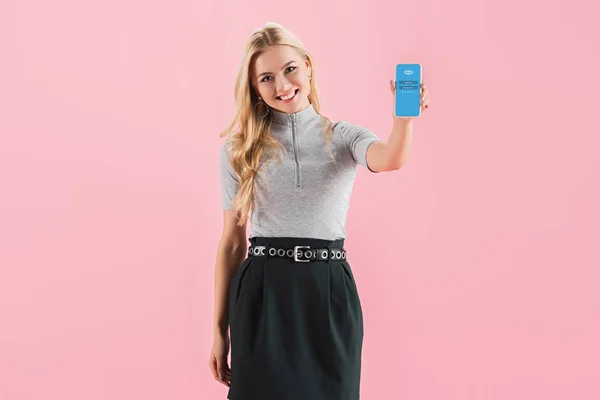 Hermosa chica sonriente que muestra el teléfono inteligente con la aplicación de Skype en la pantalla, aislado en rosa - foto de stock