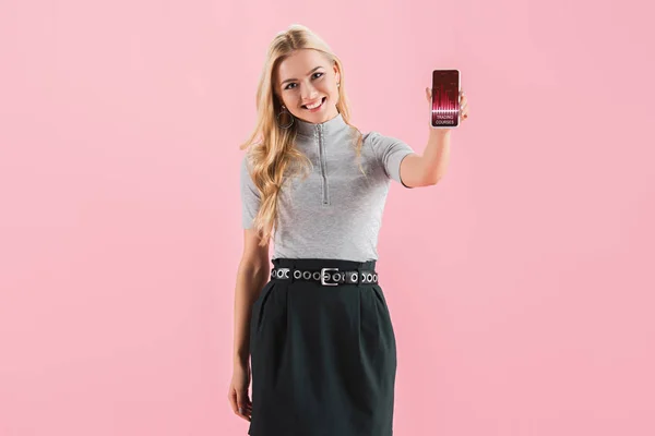 Chica rubia sonriente mostrando teléfono inteligente con cursos de comercio en la pantalla, aislado en rosa - foto de stock