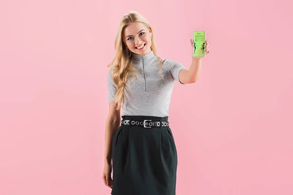Усміхнена блондинка, що показує смартфон із додатком для покупок на екрані, ізольована на рожевому — стокове фото