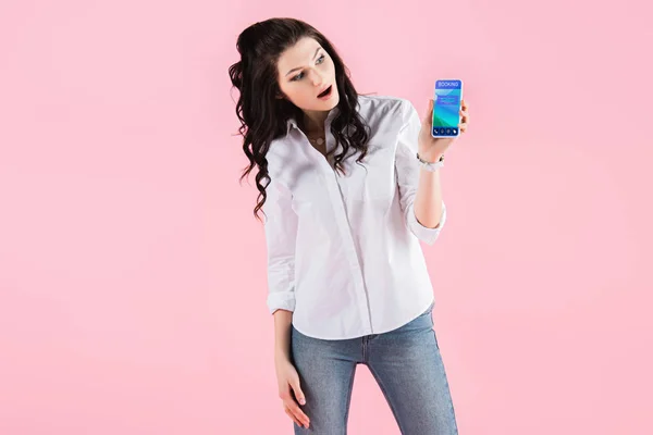 Schockierte Frau zeigt Smartphone mit Buchungs-App auf dem Bildschirm, isoliert auf rosa — Stockfoto
