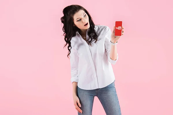 Brunette shocked girl showing smartphone with youtube app on screen, isolated on pink — Stock Photo