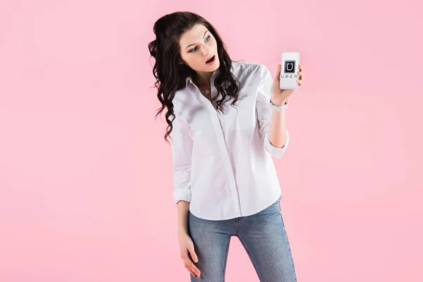 Брюнетка шокирована девушка показывает смартфон с Uber приложение на экране, изолированы на розовый — стоковое фото