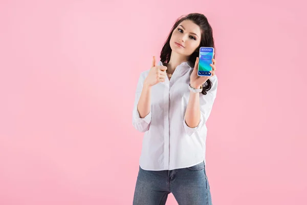 Attractive woman showing thumb up and smartphone with booking app on screen, isolated on pink — Stock Photo