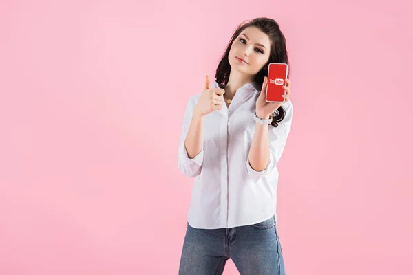 Брюнетка девушка показывает большой палец вверх и смартфон с приложением YouTube на экране, изолированные на розовый — стоковое фото