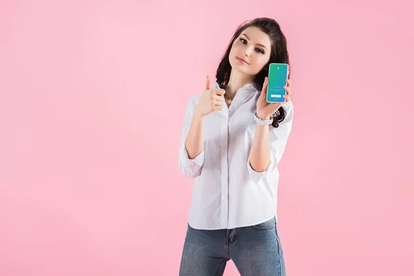 Menina morena atraente mostrando polegar para cima e smartphone com aplicativo twitter na tela, isolado em rosa — Fotografia de Stock