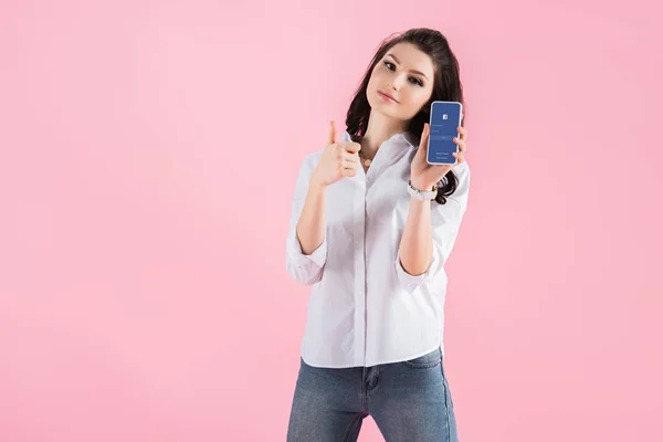 Chica atractiva que muestra el pulgar hacia arriba y el teléfono inteligente con aplicación de Facebook en la pantalla, aislado en rosa - foto de stock