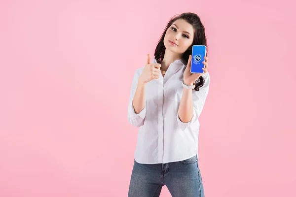 Attraktive brünette Mädchen zeigt Daumen hoch und Smartphone mit Shazam-App auf dem Bildschirm, isoliert auf rosa — Stockfoto