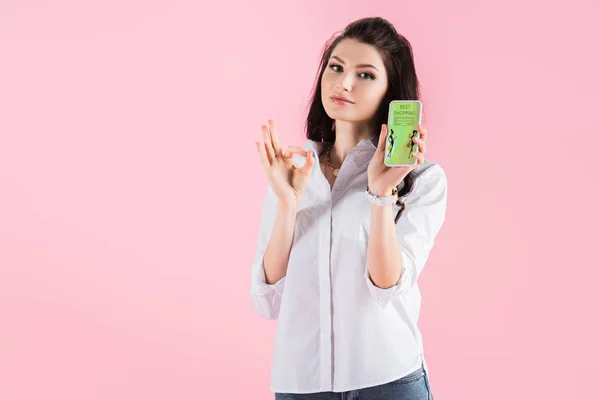 Menina morena atraente mostrando sinal ok e smartphone com aplicativo de compras na tela, isolado em rosa — Fotografia de Stock