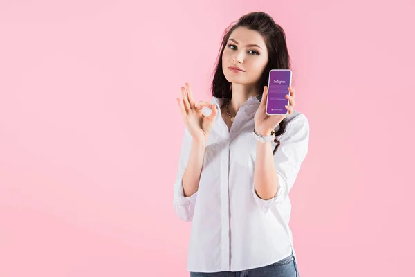 Hermosa chica morena mostrando signo de ok y teléfono inteligente con aplicación instagram en la pantalla, aislado en rosa - foto de stock