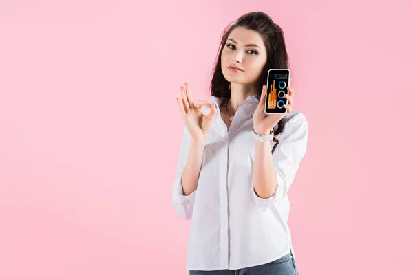 Menina morena atraente mostrando sinal ok e smartphone com aplicativo infográfico na tela, isolado em rosa — Fotografia de Stock