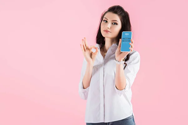 Brunette girl showing ok sign and smartphone with skype app on screen, isolated on pink — Stock Photo